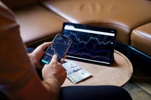 Investor reviewing stock charts and financial data on a mobile app while cross-checking with a laptop.